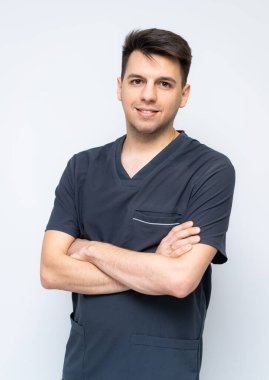 Önlüklü genç erkek sağlık çalışanı. Koyu renk önlüklü genç bir adam kollarını düz bir arkaplanda kavuşturup, profesyonelliğini gösteriyor..