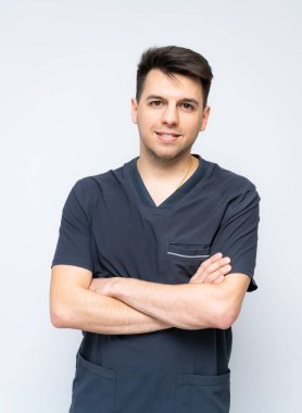 Cerrahi temizlik uzmanı pozu. Kollarını çapraz yapmış, koyu renk önlük giymiş, arka planda gülümseyen genç bir erkek doktor..