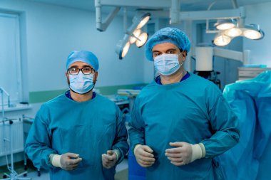 Cerrahlar ameliyata hazır. Önlüklü ve maskeli iki cerrah iyi aydınlatılmış bir hastane ameliyathanesinde tıbbi bir prosedüre hazırlanıyorlar..