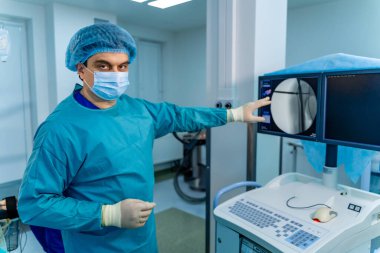 Cerrah klinik ekipmanlarını kontrol eder. Mavi önlüklü ve maskeli cerrah, ameliyata hazırlanırken parlak bir klinikte tıbbi ekipmanları teftiş eder..
