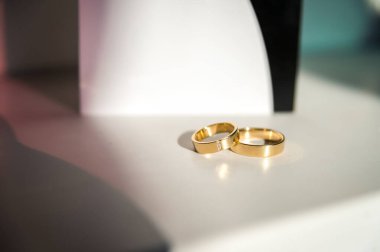 Altın alyanslar sergileniyor. İki zarif altın alyans pürüzsüz beyaz bir yüzeyde parıldıyor. Tasarımlarını vurgulamak için hafifçe aydınlatılıyor..