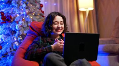 Evde sıcacık bir tatil akşamının tadını çıkarıyorum. Genç bir insan rahat bir şekilde fasulye çuvalında oturur, neşeli bir şekilde şenlikli bir akşamda dizüstü bilgisayardan bir şeyler izler..