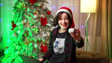 Tatil ruhu çevrimiçi toplanıyor. Noel Baba şapkası takan genç bir kadın süslenmiş bir Noel ağacının önünde arkadaşlarıyla internetten yakınlaşıyor..