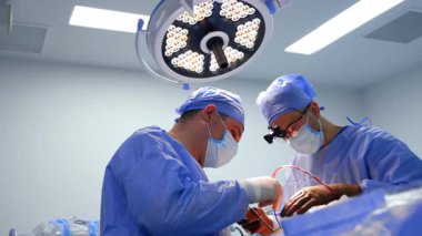 Cerrahlar ameliyat yapar. Mavi önlüklü iki cerrah iyi aydınlatılmış bir ameliyathanede cerrahi bir prosedüre odaklanıyor..