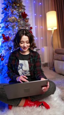Çekici gülümseyen esmer elinde dizüstü bilgisayarla yerde oturuyor. Kadınlar Noel 'de evden kumandayla çalışır. Dikey video.