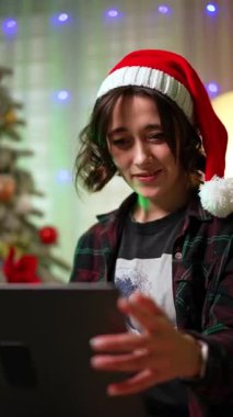 Noel Baba şapkalı, gülümseyen güzel esmer dizüstü bilgisayar tutuyor. Evden uzakta çalışan bayana alçak açılı bir bakış açısı. Noel ağacı arka planda. Dikey video.