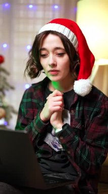 Noel Baba şapkalı güzel esmer bayan dizüstü bilgisayarda şaşırmış görünüyor. Kız mutlu bir şekilde el çırpıyor. Kapatın. Dikey video.