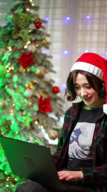 Noel Baba şapkalı mutlu beyaz kadın dizüstü bilgisayara odaklanmış. Kadın ekrana baktığında heyecanlanıyor ya da şaşırıyor. Dikey video.