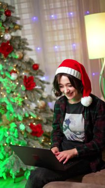 Kanepede oturup dizüstü bilgisayara bakan, Noel Baba şapkalı, gülen beyaz kız. Mutlu kadın Noel sezonunda evde çalışıyor. Dikey video.