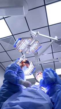 Mavi cüppeli iki doktor bir hastayı ameliyat ediyor. Cerrahların ameliyat yaparken düşük açılı görüntüsü. Dikey video.