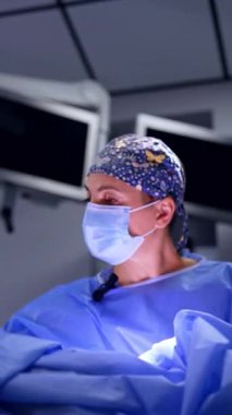 Beyaz şapkalı, maskeli ve mavi cüppeli kadın cerrah ameliyatı gerçekleştirir. Operasyona odaklanmış doktora düşük açılı bakış. Düşük açı. Dikey video.