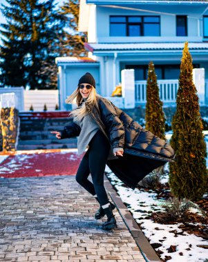 Kış gününün tadını çıkaran rahat kıyafetli bir kadın. Bir kadın, güzel bir evin yanında, şık bir kış elbisesi içinde, karlı bir patikada neşeyle yürüyor..