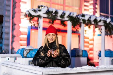 Açık hava kış pazarı. Kırmızı bereli genç bir kadın dekoratif ışıklarla çevrili açık hava kış pazarında sıcak bir içki içiyor..