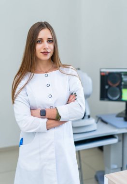 Beyaz önlüklü kendine güvenen bir kadın. Beyaz önlüklü bir kadın kollarını kavuşturup teknolojiyle donatılmış bir tıp bürosuna güveniyor..