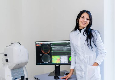 Modern klinikteki genç bayan göz doktoru. Genç bir bayan doktor güvenli bir şekilde kliniğinde duruyor ve bilgisayar ekranındaki göz taramalarını inceliyor..