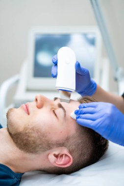 Erkekler için cilt bakımı. Modern bir klinikte cilt bakımı gören bir erkek hasta, gelişmiş ekipmanlarla yüz gençleştirme tedavisine odaklanır..