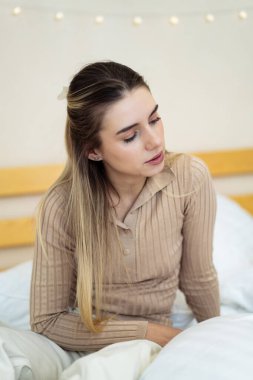 Genç bir kadın rahat bir odada yatakta dinleniyor. Genç bir kadın rahat bir odada, rahat bir odada, kabarık bir kazak giymiş, düşünceli bir şekilde yan tarafa bakıyor..