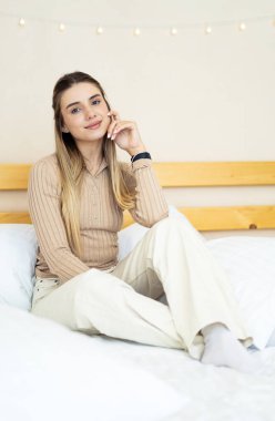 Genç bir kadın rahat bir yatakta dinleniyor. Uzun saçlı bir kadın, rahat bir şekilde bir yatakta oturuyor, rahat bir şekilde gülümsüyor, rahat giysiler içinde, boşluğunun tadını çıkarıyor..