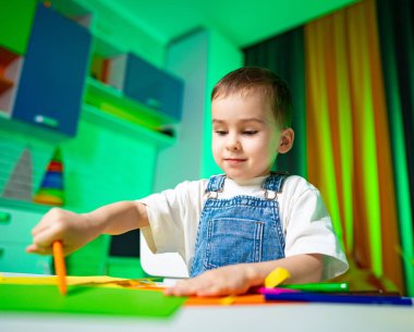 Çocuk kapalı mekan sanat projesi yapıyor. Genç bir çocuk, çeşitli materyaller kullanarak renkli bir masada canlı bir sanat projesine odaklanır..