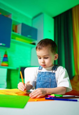 Çocuk pastel boyalarla renksiz bir oda çiziyor. Kot pantolon giymiş genç bir çocuk bir masada oturuyor, renkli, parlak renkli bir odada pastel boyalarla çizim yapıyor..