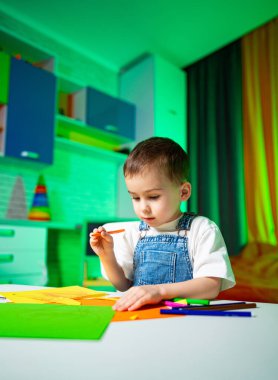 Aydınlık bir odada çocuk işi. Genç çocuk parlak ışıklı bir odada çeşitli renkli kağıtlar ve işaretler kullanarak yaratıcı faaliyetlerde bulundu..