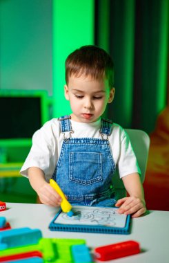 Renkli aletlerle resim yapan küçük bir çocuk. Kot pantolon giymiş genç bir çocuk, parlak renkli bir odadaki tabletin üzerindeki sarı kalemle çizime odaklanır..
