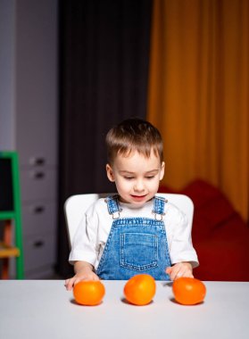 Genç çocuk evdeki masada portakalları keşfediyor. Kot pantolon giymiş genç bir çocuk evde rahat bir ortamda üç portakal yerleştirirken gülümsüyor..