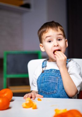 Çocuk portakal meyvesinin tadını çıkarıyor. Kot pantolonlu genç adam, masada taze portakallarla çevrili bir parça portakal tadıyor..
