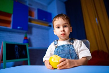 Sarı elmalı çocuk. Genç bir çocuk renkli bir odada masada oturuyor, sarı bir elma tutuyor ve doğrudan izleyiciye bakıyor..