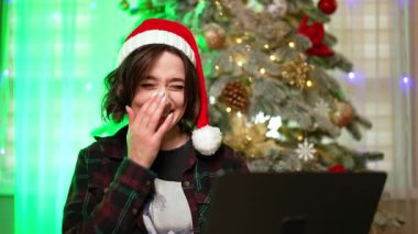 Kırmızı Noel Baba şapkalı mutlu esmer dizüstü bilgisayarına bakıyor. Kadın film izliyor ve gülüyor. Arkaplanda Noel ağacı.