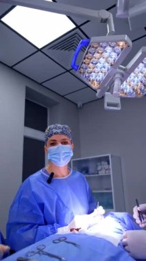 Çiçekli şapkalı üniformalı kadın cerrah ameliyat masasında kameraya konuşuyor. Modern hastanede çalışan sağlık görevlileri. Dikey video.