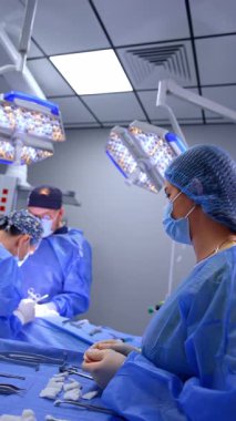 Kadın ameliyathane hemşiresi cerrahi bölümünden metal bir alet alıp cerraha pas veriyor. Arkaplanda iki cerrah ameliyat masasında çalışıyor. Dikey video.
