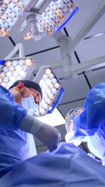 Erkek ve kadın doktorlar ameliyat yaparlar. Baş cerrah ve asistan parlak lambaların altında çalışıyorlar. Düşük açı. Dikey video.