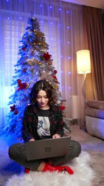 Kısa siyah kıvırcık saçlı beyaz kadın Noel ağacının yanında yerde oturuyor. Evden bilgisayarla çalışan mutlu bir bayanın portresi. Dikey video.