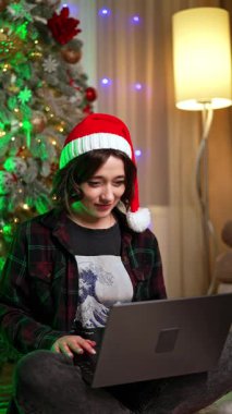 Noel Baba şapkalı esmer kız bilgisayarına hayretle bakıyor. Kadınlar Noel 'de internette çalışıyor. Dikey video.