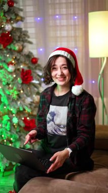 Kırmızı Noel Baba şapkalı beyaz esmer elinde dizüstü bilgisayarla kanepede oturuyor. Kız ekranı gösterir, gülümser ve el çırpardı. Noel ağacı arka planda. Dikey video.