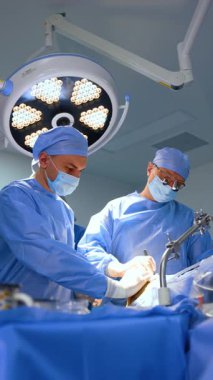 Cerrahi lensler takan profesyonel erkek cerrah ameliyat yapıyor. Erkek doktor, hastanın vücuduna destek oluyor. Dikey video.