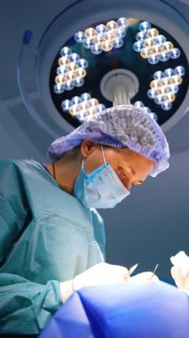 Kadın cerrah operasyonda metal aletler kullanıyor. Hastanın üzerine eğilen doktora düşük açılı bakış açısı. Dikey video.