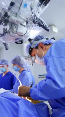 Cerrahi hoparlördeki cerrah elektrikli aletle hastanın üzerine eğildi. Plastikle kaplı bir mikroskop sıhhiyenin üstündedir. Dikey video.