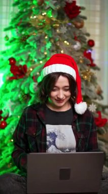 Noel Baba şapkalı güzel beyaz kadın dizüstü bilgisayarla çalışıyor. Arka planda süslenmiş Noel ağacı. Dikey video.