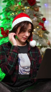 Noel Baba şapkalı esmer, bilgisayarına odaklanmış görünüyor. Noel ağacının yanındaki evden uzak bir iş. Dikey video.