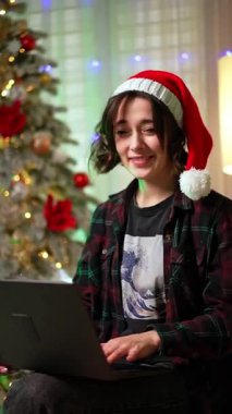 Kırmızı şapkalı güzel bayan laptopta çalışıyor. Noel zamanı evden internet üzerinden çalış. Dikey video.