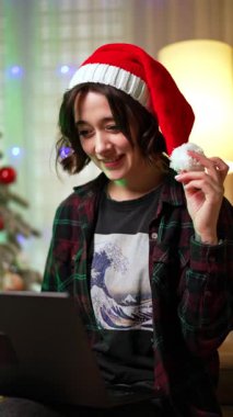 Kırmızı Noel Baba şapkalı sırıtan esmer kadın dizüstü bilgisayarına bakıyor. Mutlu düşünceli bir bayanın portresi. Noel ağacı arka planda. Dikey video.