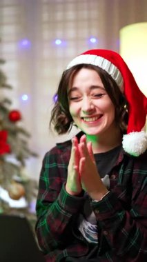 Noel Baba şapkalı mutlu, beyaz bir kız. Neşeli bayan dizüstü bilgisayara bakarak el çırpıyor. Noel ağacı arka planda. Dikey video.
