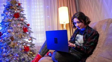 Tatillerde evde dinlenmek. Evde şenlikli bir dekorasyonla sıcak bir akşam geçiren genç bir insan, Noel ağacının yanındaki bir dizüstü bilgisayarda çalışıyor..