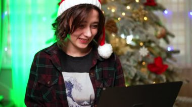 Noel ağacının yanındaki şenlikli dizüstü bilgisayar. Noel Baba şapkalı bir insan, etrafı süslenmiş bir Noel ağacı ve şenlik ışıklarıyla çevrili bir dizüstü bilgisayarda gezinmekten hoşlanır..