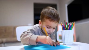 Küçük çocuk yaratıcı çizim zamanını seviyor. Evde, resim malzemeleriyle çevrili bir masada mavi kağıda renkli kalemlerle resim çizmeye odaklanmış genç bir çocuk..