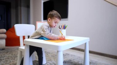 Çocuk resim çizmeyi seviyor. Küçük bir çocuk gündüz saatlerinde kapalı bir alanda küçük bir masada çizim yapmaya odaklanır..
