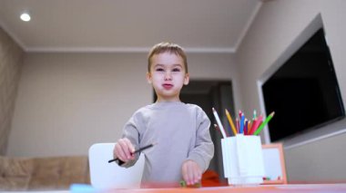 Genç çocuk evde boyamayı seviyor. Bir çocuk gururla boyama malzemelerinin olduğu bir masanın başında durur, rahat bir yerde yaratıcılık ve neşe gösterir..