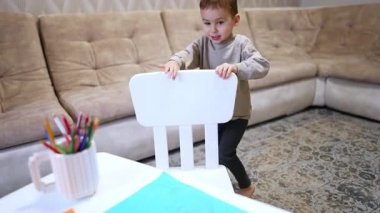 Rahat bir odada pastel boyalarla resim çizen çocuk. Genç bir çocuk, sıcak ve davetkar bir yaşam alanındaki beyaz bir masada resim yapmaya odaklanmış. Boya kalemleri düzgünce yerleştirilmiş..